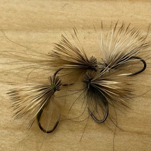 May include: Three brown and white fly fishing lures with black hooks on a wooden surface. The lures are tied with brown and white feathers and have a green accent.