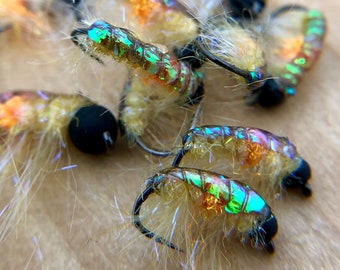 May include: Close-up of several iridescent green and orange fly fishing lures with white and brown fur. The lures are arranged on a wooden surface. The text "Jacob's Fork N.C. FLY TYING U.S." is visible in the lower right corner of the image.