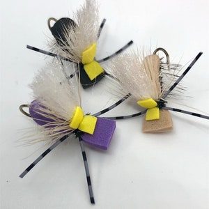 May include: Three fly fishing lures with black, tan, and purple foam bodies, white feathers, and yellow and purple accents. The lures have black and white striped legs.