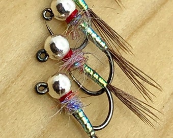 May include: Close-up of four fishing flies. Each fly has a silver bead head, a red band, and iridescent body. The hooks are black, and the tails are made of brown feathers. The text "JACOB'S FORK N.C. FLY TYING U.S." is in the lower left corner.