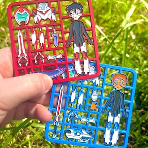 May include: Two enamel pins in the style of model kit parts. One red, one blue, each holding character and weapon designs. The characters are in black, white, and red. The background is green grass.
