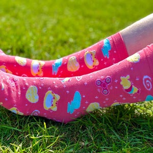 May include: A pair of bright pink socks with a playful pattern of cartoon characters, shapes, and a fidget spinner. The socks are resting on a bed of green grass, creating a vibrant and cheerful scene.