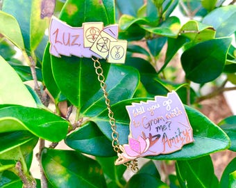 May include: A set of enamel pins featuring a light purple banner with the name "Luz" and three small cards. A second pin reads "Will you go to Grom with me? Amity" with a pink flower. The pins are connected by a gold chain.