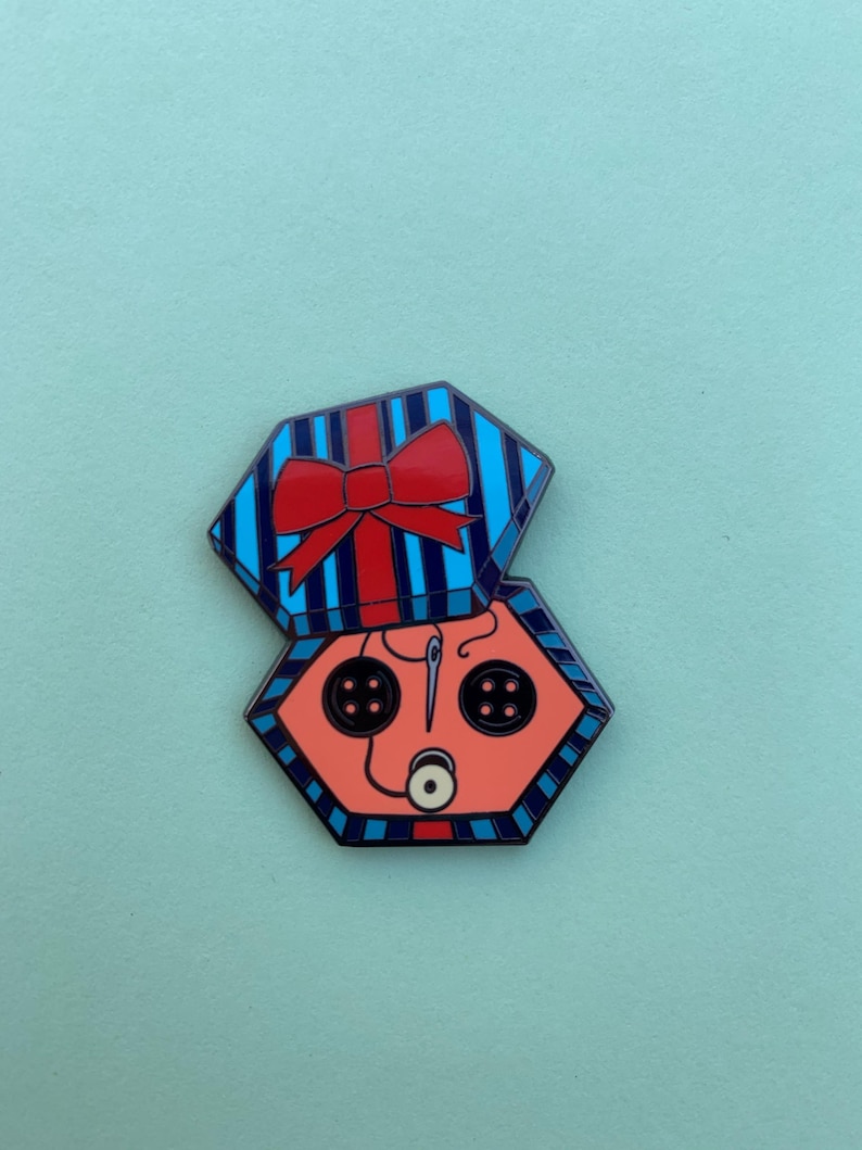 Peut inclure: Une &eacute;pingle en &eacute;mail color&eacute;e en forme de bo&icirc;te cadeau. Le dessus de la bo&icirc;te est bleu avec des rayures verticales et un n&oelig;ud rouge. &Agrave; l'int&eacute;rieur, un visage couleur p&ecirc;che avec des boutons noirs pour les yeux et une aiguille et du fil.