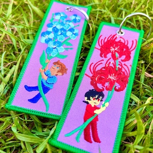 May include: Two embroidered keychains with a purple background and green trim. One features a boy holding blue flowers, the other a boy holding red flowers. Both have silver key rings.