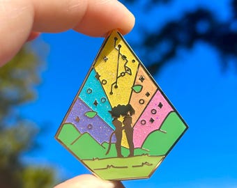 May include: A colorful enamel pin in a geometric shape, featuring a silhouette of two figures under a tree. The pin has sections of blue, purple, pink, and yellow with glitter accents. Green hills and a gold border complete the design.
