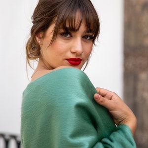 Pu&ograve; includere: Una donna con i capelli castani e la frangia indossa un maglione verde. Sta guardando oltre la spalla e sorridendo.