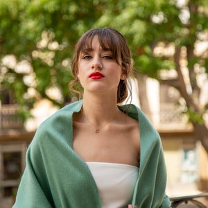 Pu&ograve; includere: Una donna con capelli castani e frangia indossa uno scialle verde sopra un top bianco senza spalline. Ha il rossetto rosso e un piercing al naso.