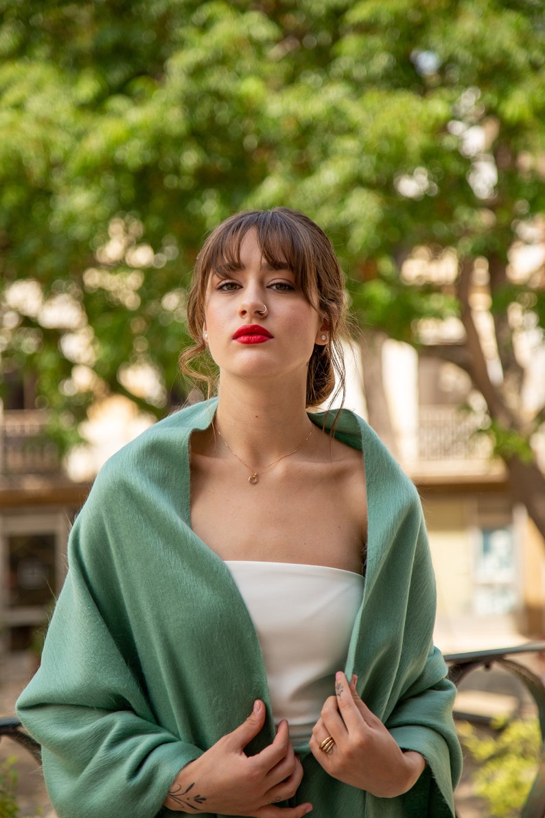 Pu&ograve; includere: Una donna con capelli castani e frangia indossa uno scialle verde sopra un top bianco senza spalline. Ha il rossetto rosso e un piercing al naso.