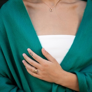 May include: A woman wearing a teal green shawl with a gold necklace and ring. The shawl is draped over her shoulders and she is holding her hand to her chest.