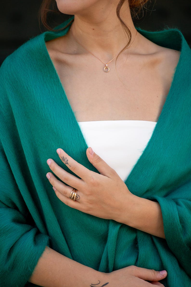 May include: A woman wearing a teal green shawl with a gold necklace and ring. The shawl is draped over her shoulders and she is holding her hand to her chest.