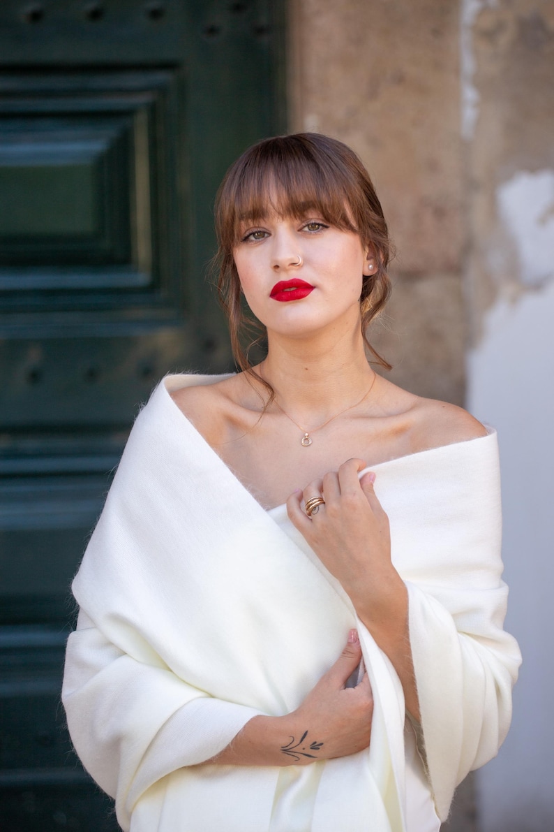 May include: A woman wearing a white shawl with a gold necklace and rings. She has red lipstick and her hair is styled with bangs.