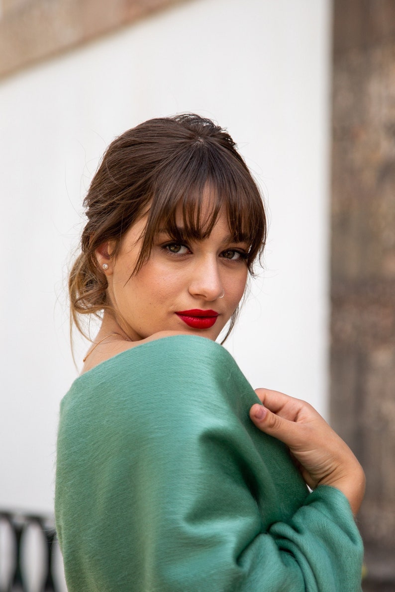 Pu&ograve; includere: Una donna con i capelli castani e la frangia indossa un maglione verde. Sta guardando oltre la spalla e sorridendo.