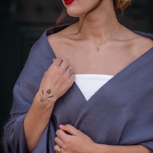 May include: A woman wearing a gray shawl with a white tank top underneath. She is wearing a gold necklace with a double circle pendant. She has a small tattoo on her wrist and is wearing gold rings on her fingers.