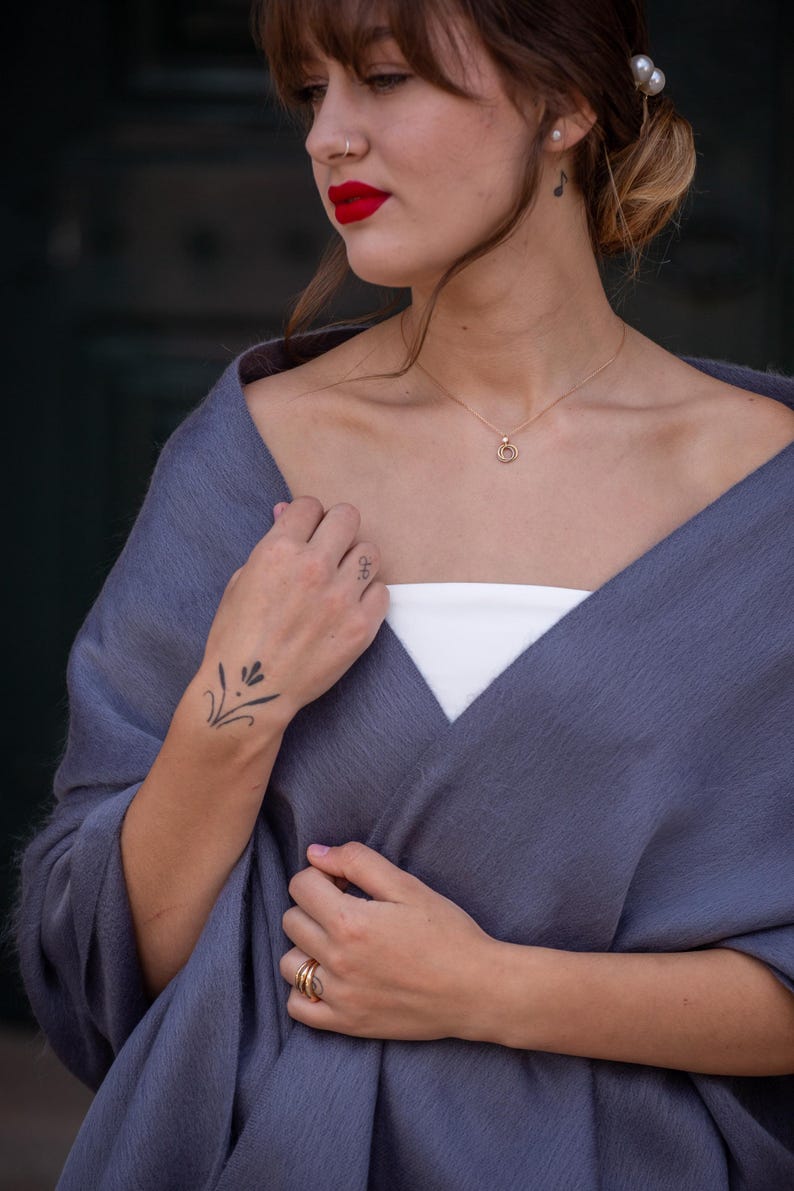 May include: A woman wearing a gray shawl with a white tank top underneath. She is wearing a gold necklace with a double circle pendant. She has a small tattoo on her wrist and is wearing gold rings on her fingers.