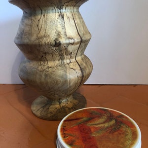 Puede incluir: Un jarrón de madera torneada con un patrón de vetas marrón claro y marrón oscuro. El jarrón tiene una base ancha, una sección media bulbosa y una parte superior acampanada. Una pila de posavasos redondos con un diseño de palmera está al lado del jarrón.