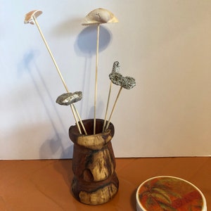 May include: A wooden vase holds several decorative stems topped with seashells and stones. A stack of four coasters with a tropical palm tree design sits to the right. The vase and coasters are on a brown surface.
