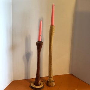 May include: Two wooden candle holders with pink candles. One is dark brown with a flared shape and sits on a wooden slice. The other is light brown and tall. The candles are the same color.