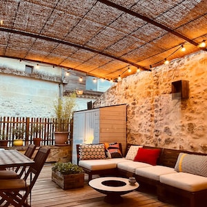 May include: A white sectional sofa with colorful pillows sits on a wooden deck under a pergola with string lights. The sofa is facing a stone wall with a small wooden shed to the left.