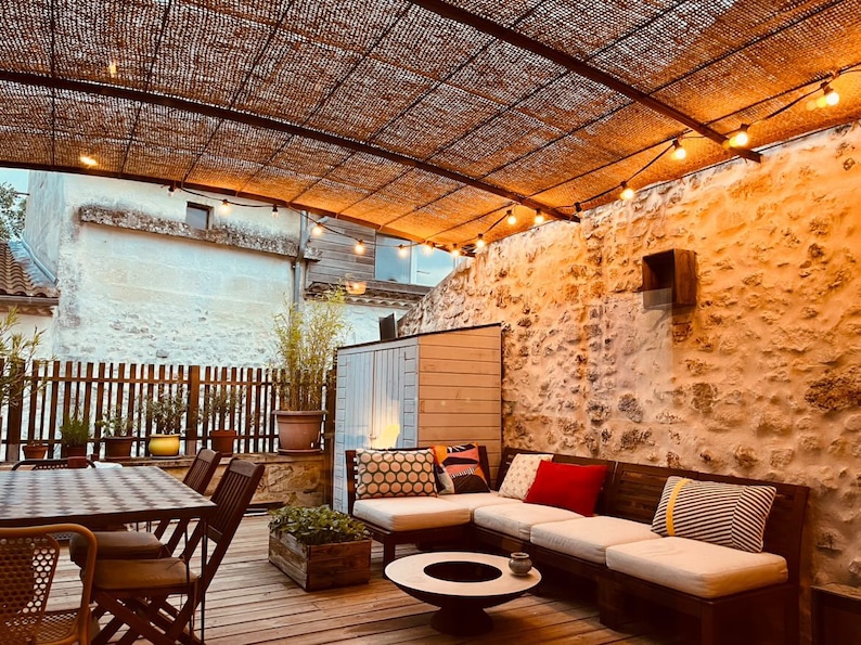 May include: A white sectional sofa with colorful pillows sits on a wooden deck under a pergola with string lights. The sofa is facing a stone wall with a small wooden shed to the left.