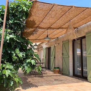 May include: A patio with a brown woven shade structure over a tiled floor. The patio has a table and chairs, and a green fig tree to the left. The house has green shutters and a sliding glass door.