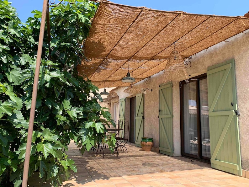 May include: A patio with a brown woven shade structure over a tiled floor. The patio has a table and chairs, and a green fig tree to the left. The house has green shutters and a sliding glass door.