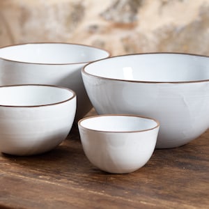 Peut inclure: Quatre bols en céramique blanche avec des bords bruns, disposés par ordre de taille du plus grand au plus petit sur une surface en bois.