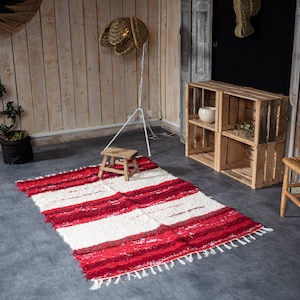 May include: A red and white striped rug with fringe on the edges. The rug is laid out on a gray concrete floor in a room with wooden walls and a wooden stool.