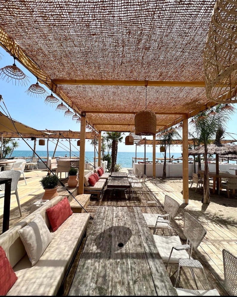 May include: A wooden table with a weathered finish sits under a thatched roof canopy.  The table is surrounded by white wicker chairs and a white couch with red pillows.  The ocean is visible in the background.