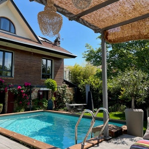 May include: A swimming pool with a wooden deck and a pergola covered in a brown woven material. The pool is surrounded by lush greenery and a brick house with large windows.