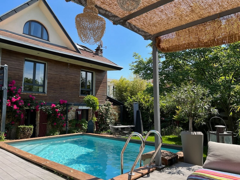 May include: A swimming pool with a wooden deck and a pergola covered in a brown woven material. The pool is surrounded by lush greenery and a brick house with large windows.