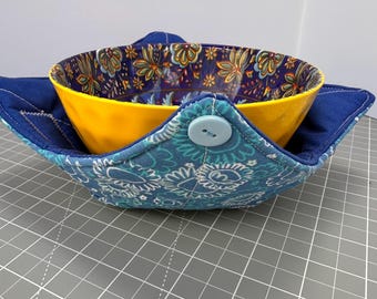 Könnte beinhalten: Eine gelbe Keramikschüssel befindet sich in einer blauen und türkisfarbenen Stoffschüssel-Hülle. Die Hülle hat ein Blumenmuster und wird mit einem Knopf befestigt. Das Innere der Schüssel hat ein Blumenmuster in Blau, Gelb und Grün.