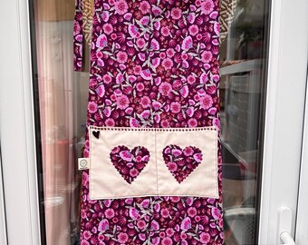 Könnte beinhalten: Eine blumige Schürze mit tiefviolettem Hintergrund und rosa Blumen. Die Schürze hat eine cremefarbene Tasche mit zwei gestickten Herzformen. Die Schürze hängt an einem Kleiderbügel.