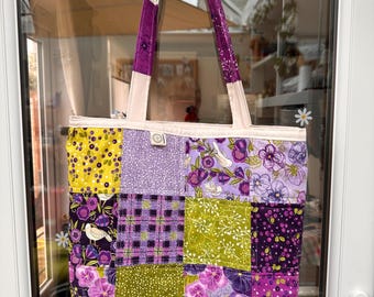 Könnte beinhalten: Eine gesteppte Tragetasche mit einem Patchwork-Design aus verschiedenen Stoffquadraten in Lila-, Lavendel-, Limettengrün- und Weißtönen. Die Muster umfassen Blumendrucke, Beeren und Karos. Die Tasche verfügt über zwei lange Schulterriemen und ein Etikett.