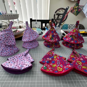 May include: A collection of triangular fabric pot holders featuring two distinct patterns. One set displays a light purple background with small dark blue and pink leaf motifs, while the other set features a vibrant red background with floral designs.