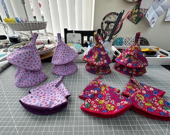 Könnte beinhalten: Eine Sammlung dreieckiger Stofftopflappen mit zwei verschiedenen Mustern. Ein Set zeigt einen hellvioletten Hintergrund mit kleinen dunkelblauen und rosafarbenen Blattmotiven, während das andere Set einen leuchtend roten Hintergrund mit Blumenmustern aufweist.