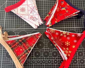 Könnte beinhalten: Vier Sets aus dreieckigen Stoffwimpeln. Ein Set zeigt ein weiß-rotes Schneeflockenmuster mit roter Einfassung, ein weiteres einen roten Hintergrund mit bunten geometrischen Hirschen, ein drittes ein rotes Schottenmuster mit goldenen Sternen und das letzte goldene Schneeflocken.