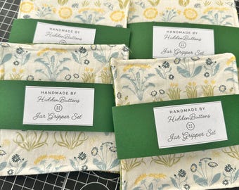 Könnte beinhalten: Ein Set aus vier quadratischen Glasöffnern aus Stoff mit einem Blumenmuster in Gelb, Blau und Grün auf weißem Grund. Jeder Glasöffner ist mit einer grünen Papierbanderole und einem weißen Etikett mit der Aufschrift "HANDMADE BY Hidden Buttons Jar Gripper Set" versehen.