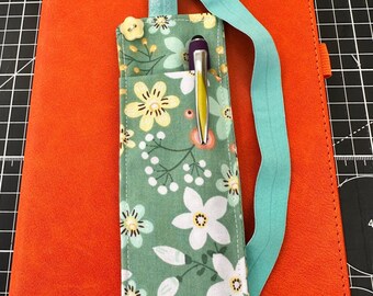 Könnte beinhalten: Ein grüner Stiftehalter aus Stoff mit weiß-gelbem Blumenmuster, einem hellblauen Gummiband und einem kleinen cremefarbenen blumenförmigen Knopf. Der Text 5. White flowers ist oben links auf dem orangefarbenen Notizbuch aufgedruckt.