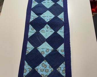 Könnte beinhalten: Gesteppter Tischläufer mit einem Rautenmuster in Blautönen aus Jeansstoff und hellblauem Stoff. Die Rauten sind von einem dunkelblauen Stoff umrandet. Der Läufer ist ca. 30 cm breit und 91 cm lang.