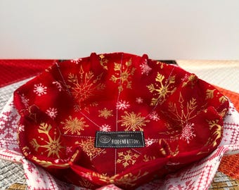 Könnte beinhalten: Ein roter Stoffnapf mit goldenen Schneeflocken und einer weiß-roten Schneeflockenmuster-Außenseite. Die Schüssel hat ein Etikett mit der Aufschrift "HANDMADE BY HIDDENBUTTONS". Die Schüssel wird wahrscheinlich zum Servieren oder Aufbewahren verwendet.