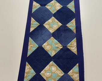 Könnte beinhalten: Rechteckiger Tischläufer mit marineblauem Rand und sich wiederholendem Rautenmuster. Die Rauten wechseln sich ab zwischen einfarbigem Marineblau und einem beigen Stoff mit blau-weißem Blumenmuster. Der Läufer ist ca. 30 cm breit und 91 cm lang.