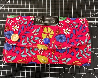 Könnte beinhalten: Eine rechteckige Stoffgeldbörse mit einem leuchtend pinken Hintergrund und einem mehrfarbigen Blumen- und Blattmuster. Sie verfügt über ein schwarzes Etikett mit der Aufschrift "HANDMADE BY HIDDENBUTTONS", einen hellen Holzknopf und einen kleinen blauen Knopf.