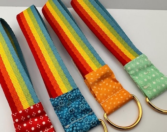 Könnte beinhalten: Vier farbenfrohe Schlüsselanhänger mit Regenbogen-Streifen. Jeder Anhänger hat einen anders gemusterten Stoffakzent in Rot mit weißen Punkten, blau floral, orange mit weißen Punkten und grün mit weißen Punkten. Goldfarbene Metallringe sind befestigt.