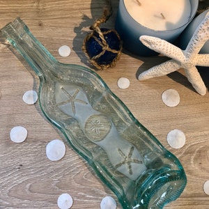 May include: A clear glass bottle with a textured surface, shaped like a serving dish. The bottle is decorated with etched images of starfish and sand dollars. The bottle is sitting on a wooden surface with white sand dollars scattered around it.