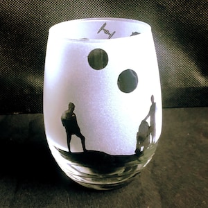 May include: A frosted glass wine glass with a black silhouette of two figures and a spaceship against a white background. The figures are standing on a black hill with two black circles above them.