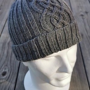 May include: A gray knitted beanie with a cable knit pattern. The beanie is on a mannequin head.