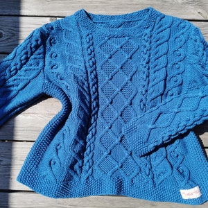 Può includere: Un maglione blu lavorato a maglia con un motivo a treccia. Il maglione ha un collo rotondo e maniche lunghe.