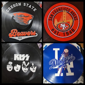 Può includere: Quattro dischi in vinile con design diversi. Il primo disco è nero con testo bianco che dice "Oregon State Beavers 1868" e un logo di castoro rosso e nero. Il secondo disco è rosso con testo bianco che dice "San Francisco 49ers est. 1946" e un design di ponte dorato. Il terzo disco è nero con testo bianco che dice "KISS". Il quarto disco è blu con testo bianco che dice "Los Angeles Dodgers" e un logo di baseball rosso e bianco.