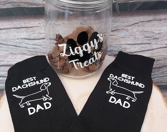 Puede incluir: Un tarro de vidrio con tapa de metal y una etiqueta que dice "Ziggy's Treats". El tarro está lleno de golosinas para perros. Dos calcetines negros con siluetas de dachshund blancas y el texto "Best Dachshund Dad" están sobre una superficie de madera.